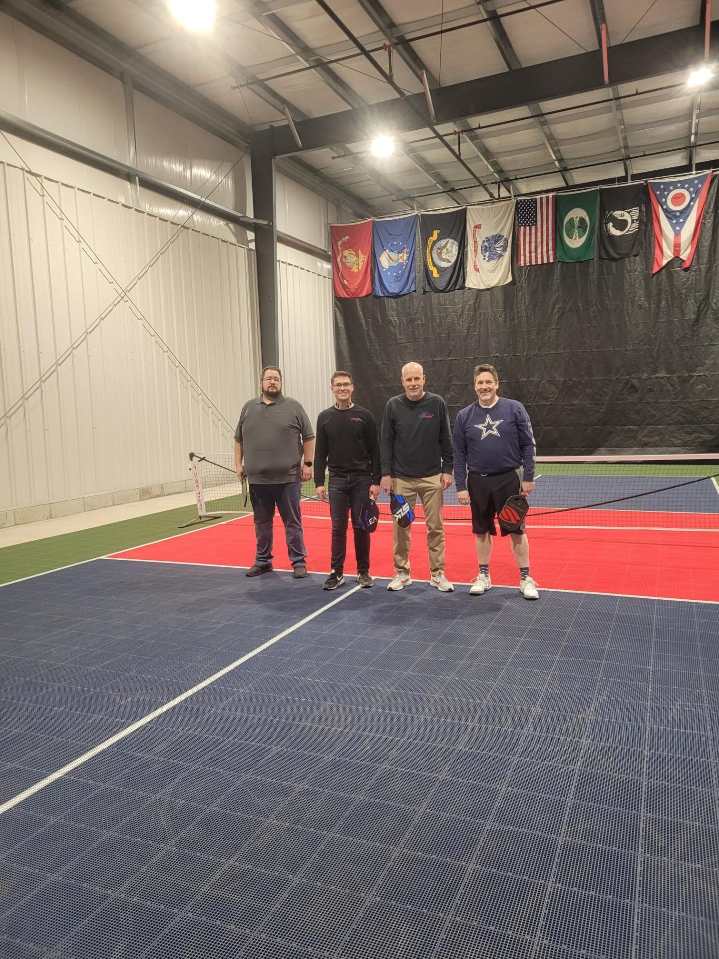 Four team members posing on the CMF pickleball court with military flags